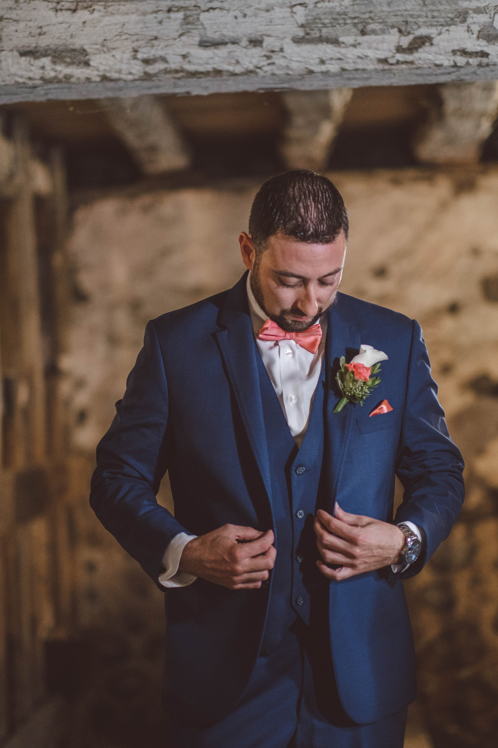 Groom buttons up his suit before his wedding.  Wearing a blue suit with a pink bowtie, standing in a rustic location of a barn.