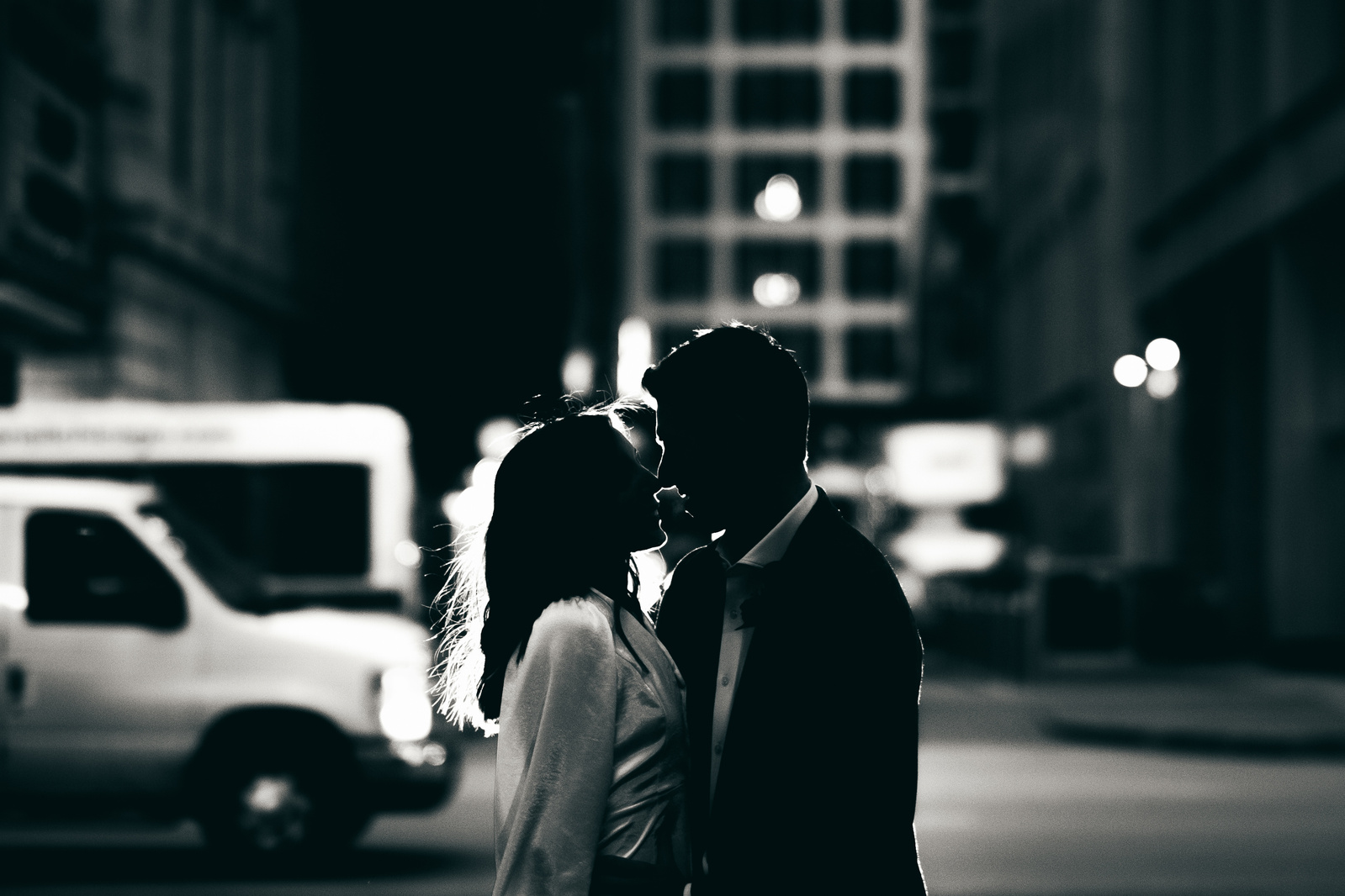 A newlywed couple kisses with city lights and passing traffic behind them after they were married in a courtroom in Chicago, Illinois.  The photo is in black and white and is a silhouette shot of the bride and groom.