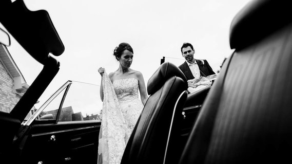 Wedding couple gets into convertible classic car on their wedding day. Bride is holding dress in her right hand, looking down into the car and the groom is in the background, helping the bride into the car.