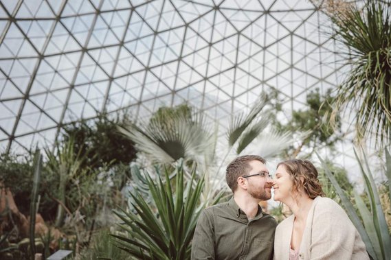 couple snuggles at the milwaukee domes for an engagement session.  nose to nose, eyes closed, they sit in a playful position under the glass dome and amongst the desert foliage.