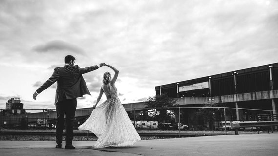 Wedding couple dances in an alley on their wedding day.  Groom is spinning bride, he is wearing a tuxedo and she is wearing a white dress.  Photography is in black and white.