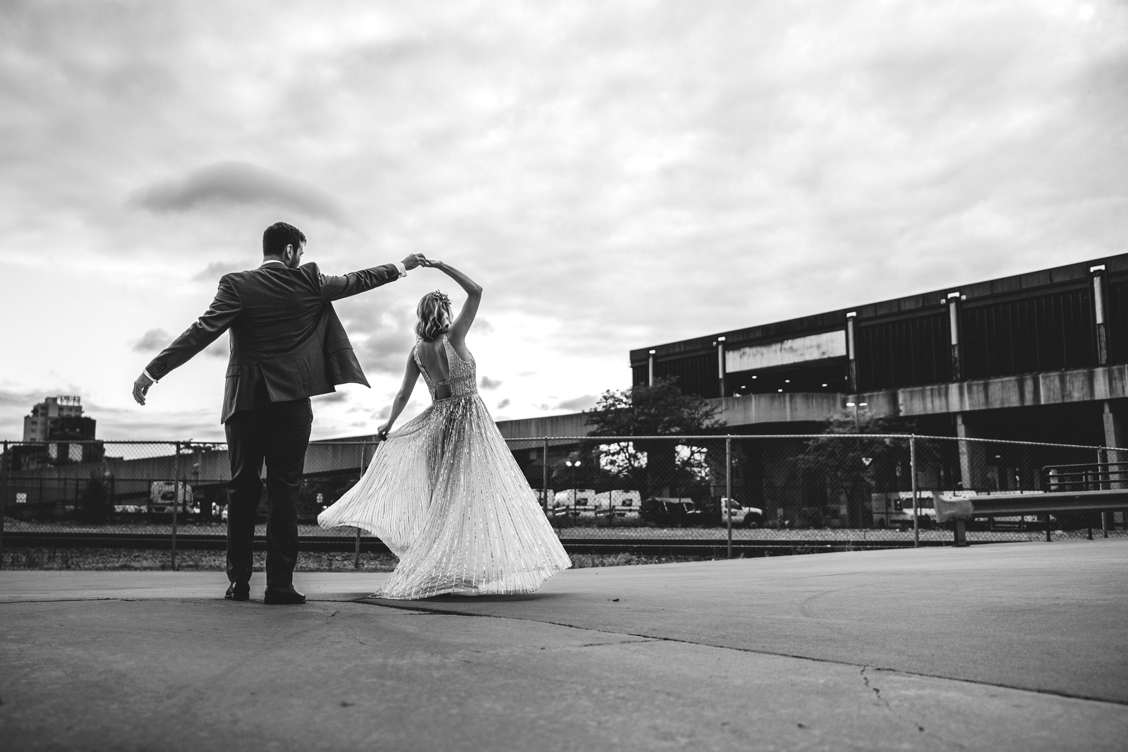 black and white photograph of couple dancing outside under a cloudy sky during their wedding.  Urban environment, with parking structure as the backdrop to groom twirling the bride in epic fashion.