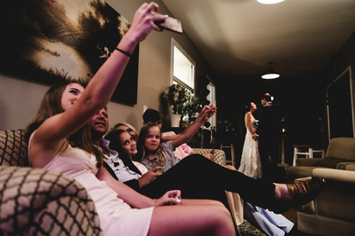 candid photograph of family taking a group selfie on a couch while bride and groom dance alone on the dance floor in the background.