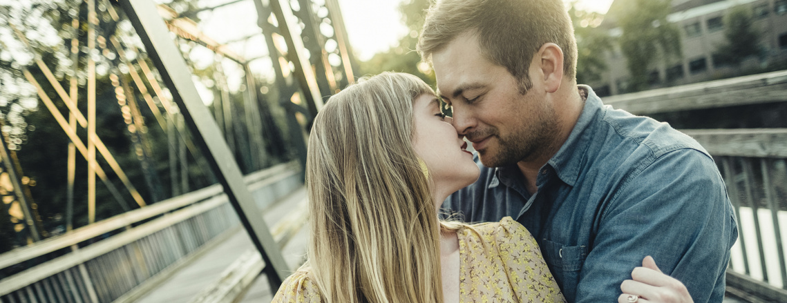 A couple embraces on a bridge during their engagement shoot, nearly kissing.  The woman is wearing a yellow flower dress and the man is wearing a blue shirt.  They are good looking, and you can see the woman's engagement ring on her left hand.