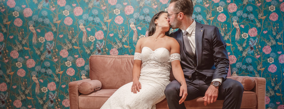 wedding couple sits on pink couch kissing in front of aqua colored flower and bird wallpaper.  wedding is at city view lofts in chicago, illinois.