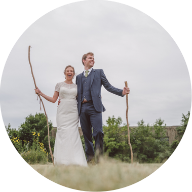 couple standing in a field in front of trees holding sticks.  they are smiling and laughing, and bride is wearing a wedding dress and groom is wearing a suit.  Milwaukee Wedding at the Urban Ecology Center, taken by Patrick Betcher of Betcher Photo.