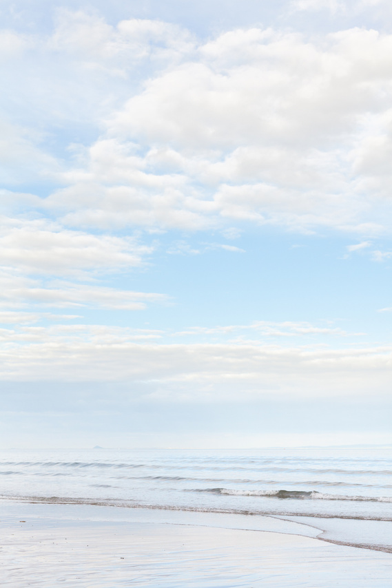 Pale blue seascape, looking into calm waves on a beach with blue sky and white clouds. 