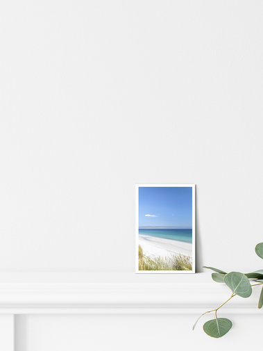 Portrait photographic postcard print. White sandy beach landscape, with turquoise sea and blue sky. Shown on a mantelpiece with light coastal decor. A6 with a 5 mm white border.