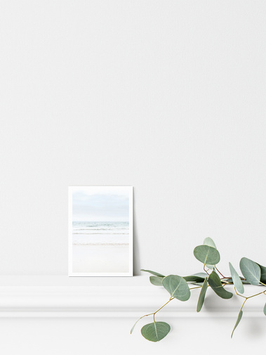 Portrait photographic postcard print. Pale blue seascape, looking into waves from a sandy beach. On a mantelpiece with light coastal decor. A6 with a 5 mm white border.