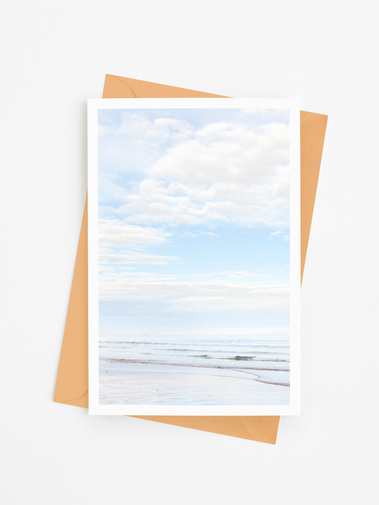 Portrait photographic beach greetings card. Pale blue seascape, looking into calm waves on a beach with blue sky and white clouds. A6 card with a 5 mm white border.