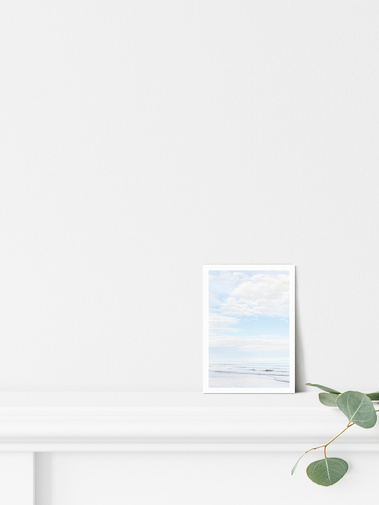 Portrait photographic beach postcard print. Pale blue seascape, looking into calm waves on a beach with blue sky and white clouds. Shown on a mantlepiece with light coastal decor. A6 postcard with a 5 mm white border.