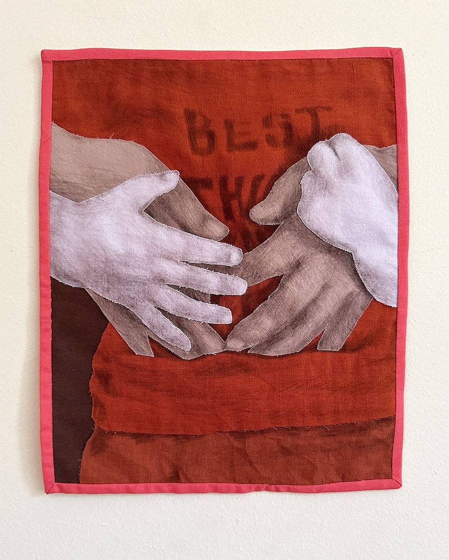 A small wall hanging of a child hugging an adult's hands to their chest. Only the two pairs of hands and clothes underneath are visible. The layers are cut from lilac and orange fabric with details drawn on with brown pencil with a bright pink border