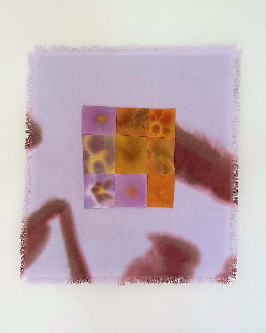 A small wall hanging with frayed edges. The  central motif is a patchwork of nine squares that each depict an abstracted flower in orange or purple. The background is pale lilac with brown gestural marks.