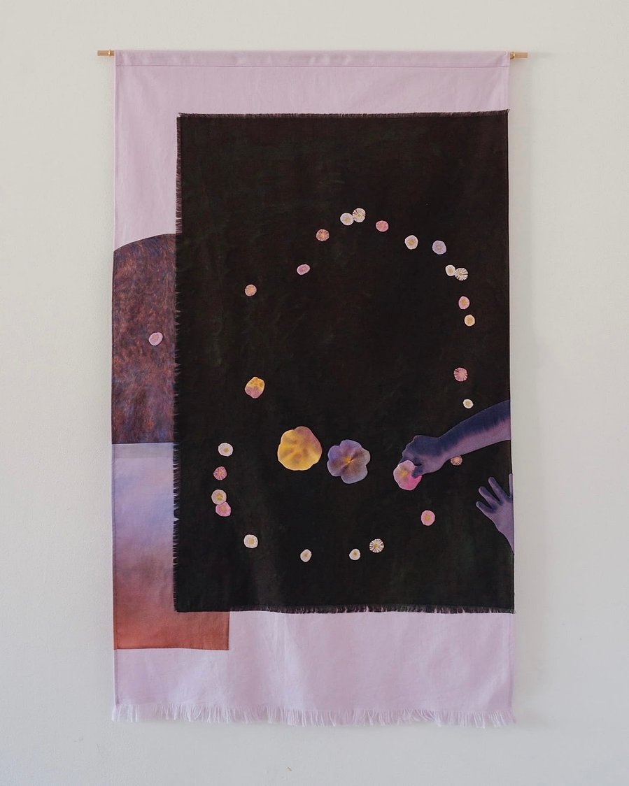 A wall hanging with collaged layers of painted appliquéd fabric. The main motif is a child's hands reaching in from the side arranging flowers in a constellation-like pattern onto a dark green-black background.