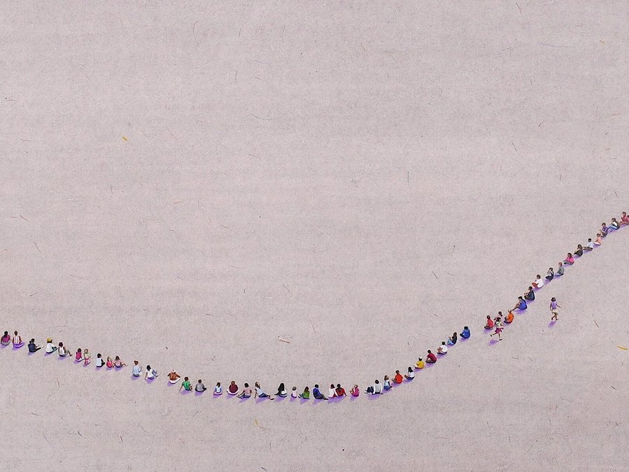A colour pencil drawing of tiny people against a vast grey background wash. The figures are seated in an enormous circle with hundreds of participants playing 'duck, duck, goose’