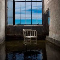 Abandoned room in Packard Plant, Detroit, MI. A white chair sits in a puddle, with a subtle reflection. Deep blue skies are visible through the window, adding a sense of serenity to this captivating scene