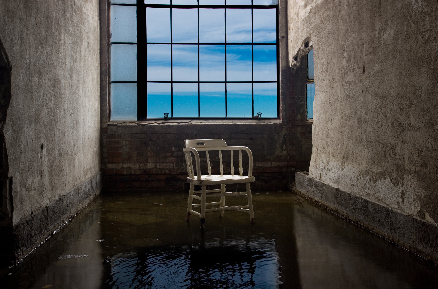 Abandoned room in Packard Plant, Detroit, MI. A white chair sits in a puddle, with a subtle reflection. Deep blue skies are visible through the window, adding a sense of serenity to this captivating scene