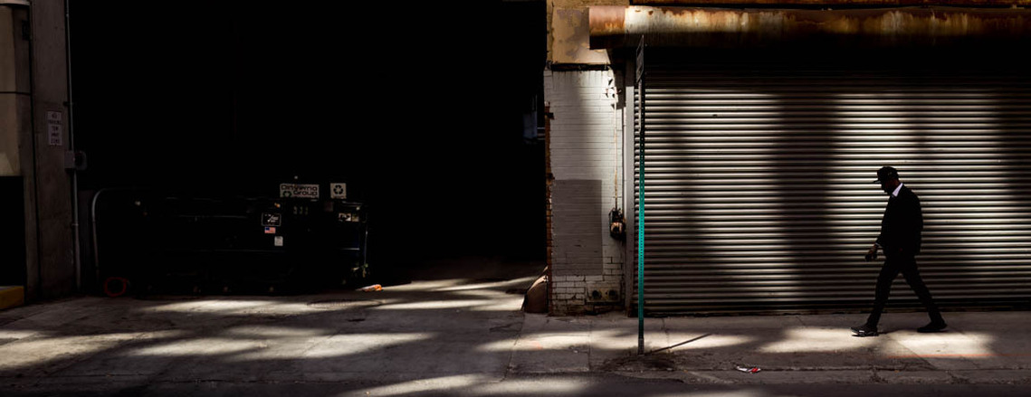 In Detroit's vibrant streets, a stylish man walks through patches of light, casting captivating shapes on the ground and walls. Silhouetted in the brilliance, he exudes confidence, donning fashionable attire and a sleek hat. A dynamic blend of light, fash