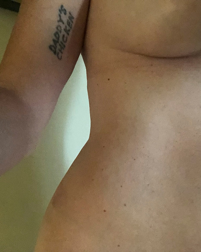 A woman with a tattoo that reads "Daddy's Chicken" on her right bicep takes a selfie of her mid section standing nude in an apartment in Los Angeles, CA. The underside of her breast and, waist and curvy hips are displayed.