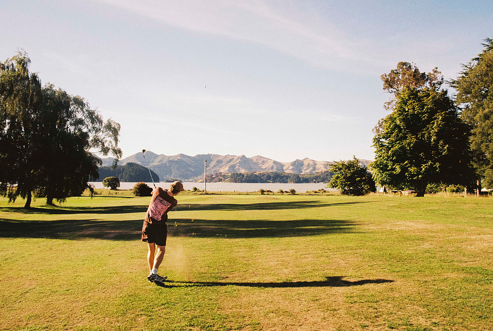 A wide-angle 35mm film shot of a golfer hitting the ball toward the green, with a scenic landscape in the background. Captured on the Canon AE-1 Program with the Tokina 28mm f/2.8 FD lens.