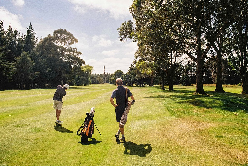 A wide-angle 35mm film image of a golfer taking a shot while another watches beside him. The Canon AE-1 Program captures the scene with natural contrast and rich film tones."