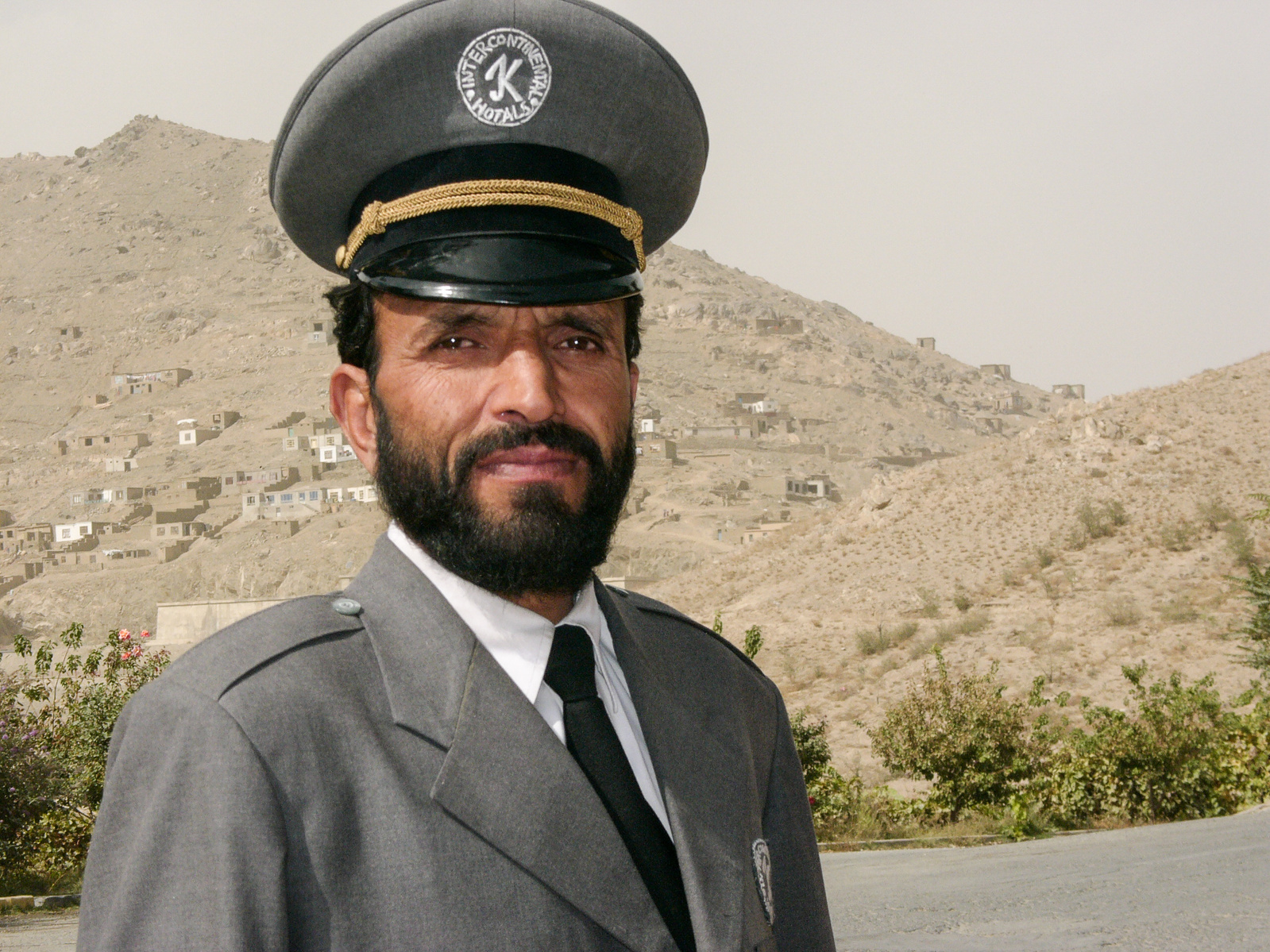 A bearded man in a grey uniform, the gatekeeper of the Intercontinental Hotel in Kabul, Afghanistan, in front of the arid landscape around the hotel.
