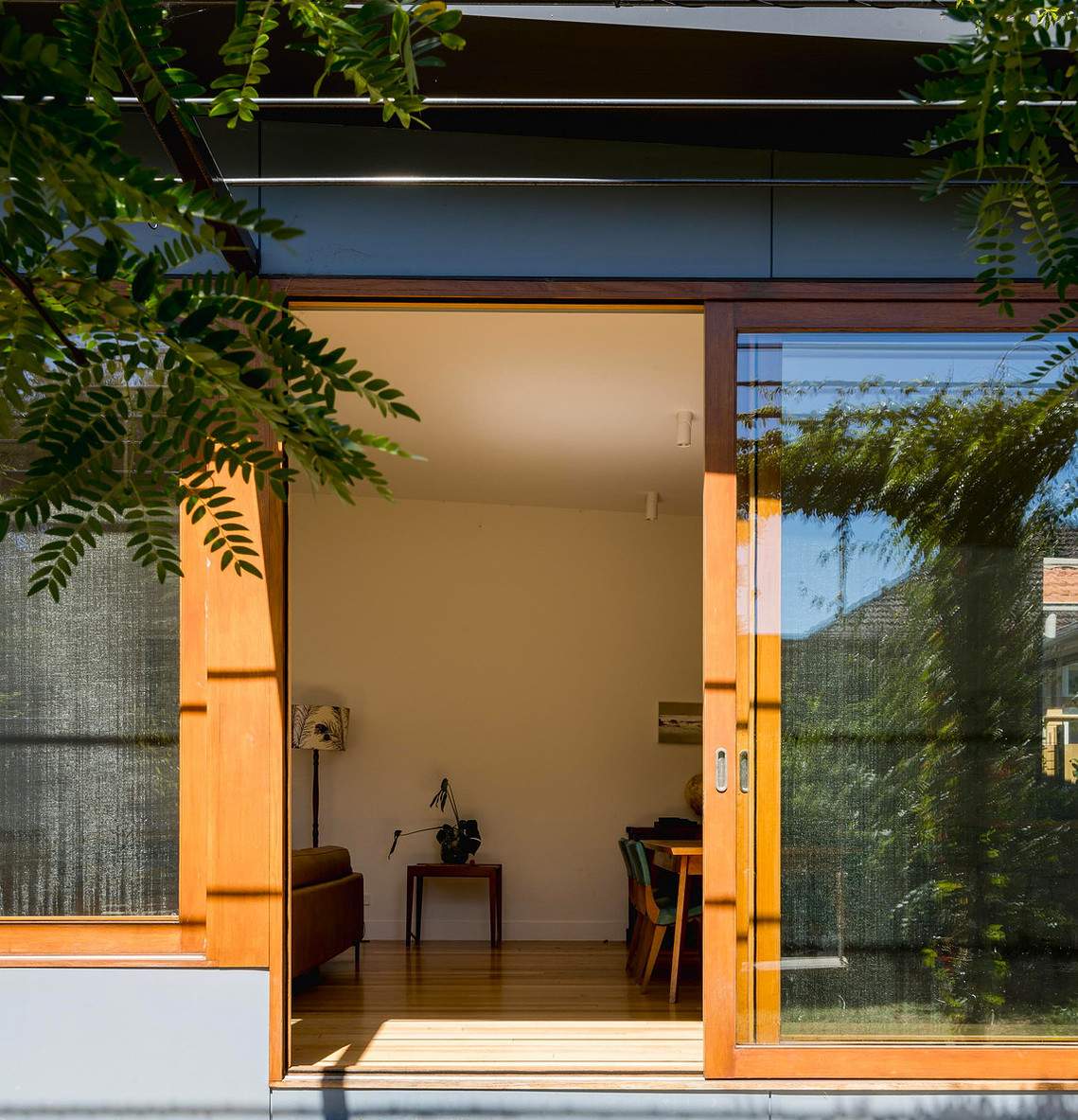Large timber doors open up from outside to a north facing living space. Plants are seen reflected in the glass.