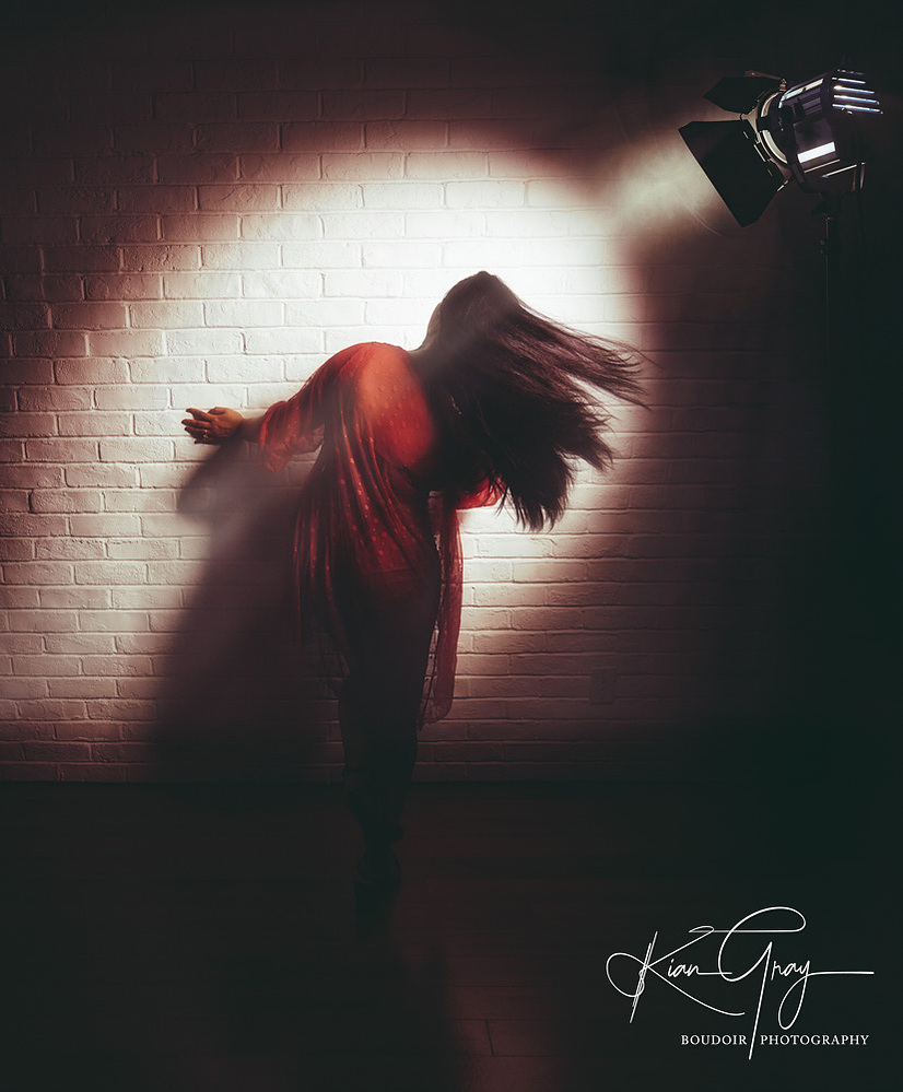 A striking boudoir photo showcasing movement and mystery with dramatic lighting, a flowing red robe, and cinematic shadows against a white brick wall.