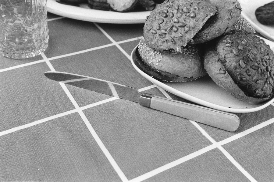 Black and white photograph of a knife that reflects part of the table cloth and a few bread buns