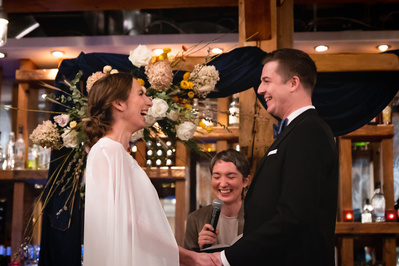 A wedding officiant stands behind couple in wedding ceremony with all three laughing.