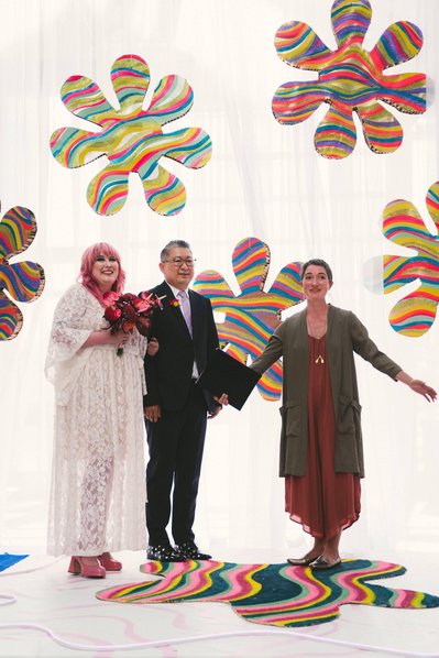 A bride with pink hair and groom in a dark suit stand beside a wedding officiant on a white dais with brightly coloured 1970s style  flower cutouts hanging around them.