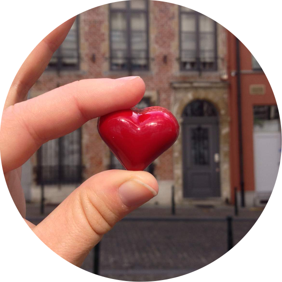 A red chocolate heart is held between thumb and forefinger against a picturesque street.