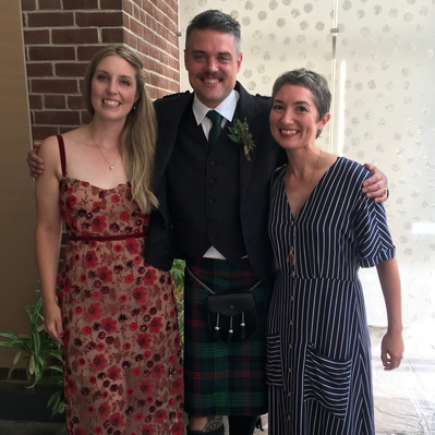 A wedding officiant poses and smiles with groom dressed a green tartan kilt and bride dressed in a red dress.