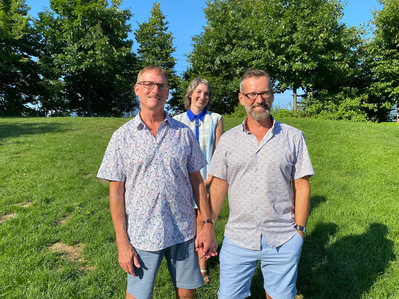 Two grooms and a wedding officiant stand together smiling with a bright blue sky and trees in background.