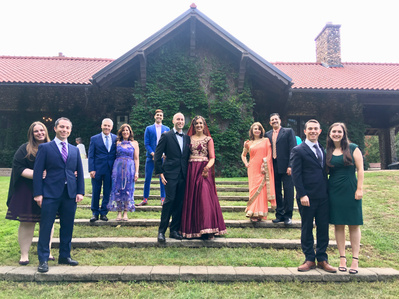 A colourfully dressed group of wedding guests gathered around a bride and groom on a hill in front of a building.