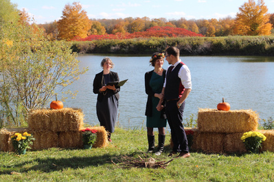 A wedding officiant standing off to the side of couple being married in front of a lake on a vibrant fall day.
