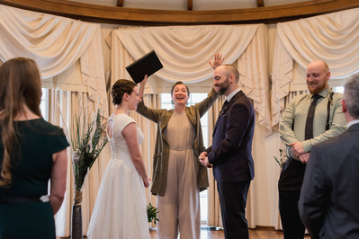 A bride and groom standing facing each other in front of wedding officiant with arms raised ecstatically behind them.