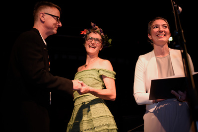 A wedding officiant speaks into a mic while a couple dressed in black suit and green dress look on grinning.