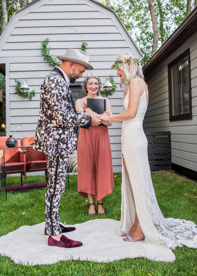 A bride and groom exchange rings in a backyard ceremony while a wedding officiant looks on.
