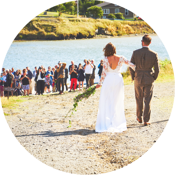 A woman in a white wedding dress and man in a khaki suit look back on a group of guests at a wedding against the backdrop of a body of water and a hill climbing behind it.