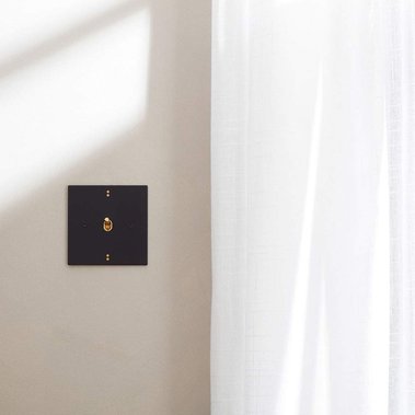 Product photography of a black designer light switch on a beige wall with white linen curtains.