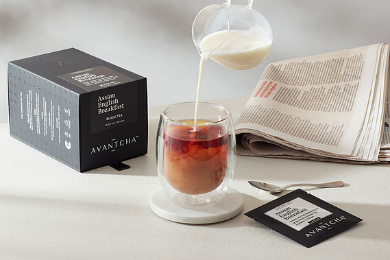 Tea product photography.  A cup of Assam Breakfast tea with milk being poured in.  Tabletop scene with a newspaper and product packaging on a neutral tabletop