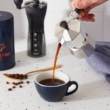 Coffee product photography, coffee being poured from a stovetop pot into a cup on a white tabletop