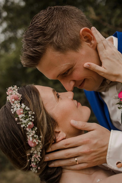 Brautpaar nehmen Gesichter in die Hand vor dem Kuss und sie hat einen zarten weiss rosa Blumenkranz in ihren Haaren