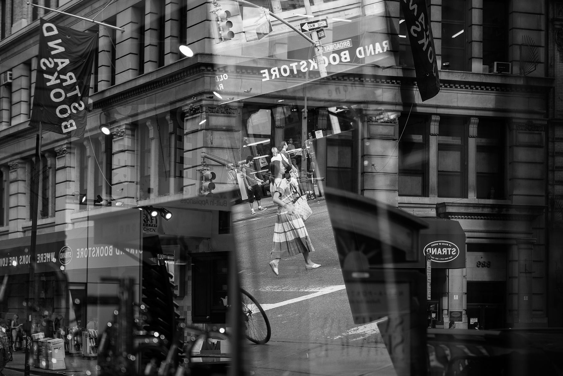A black and white street photograph of a pedestrian in an intersection. The image has multiple reflections from windows and mirrors