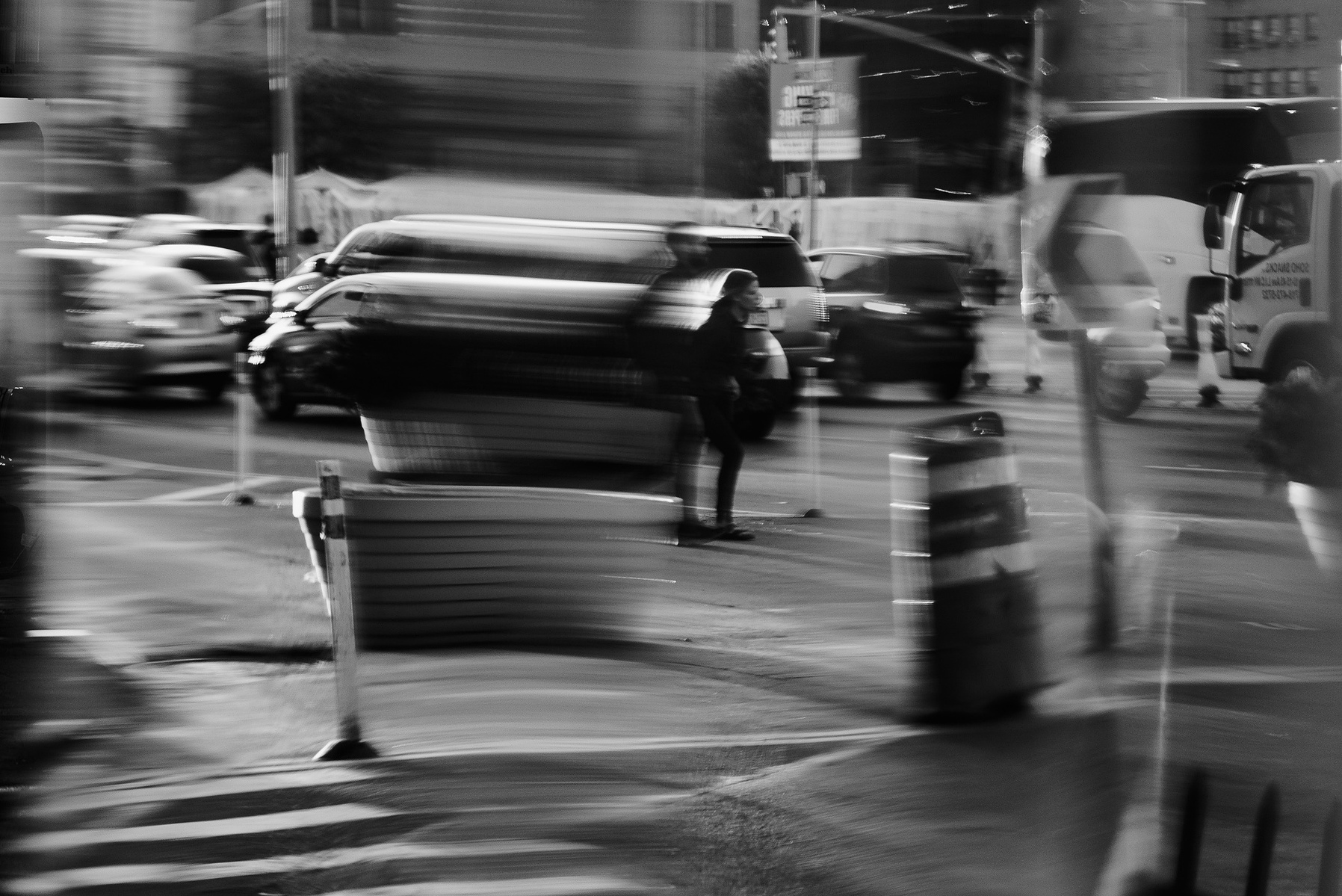 A black and white street photograph of two people crossing the street. The image was taken in the reflection of a piece of metal, and has several distortions