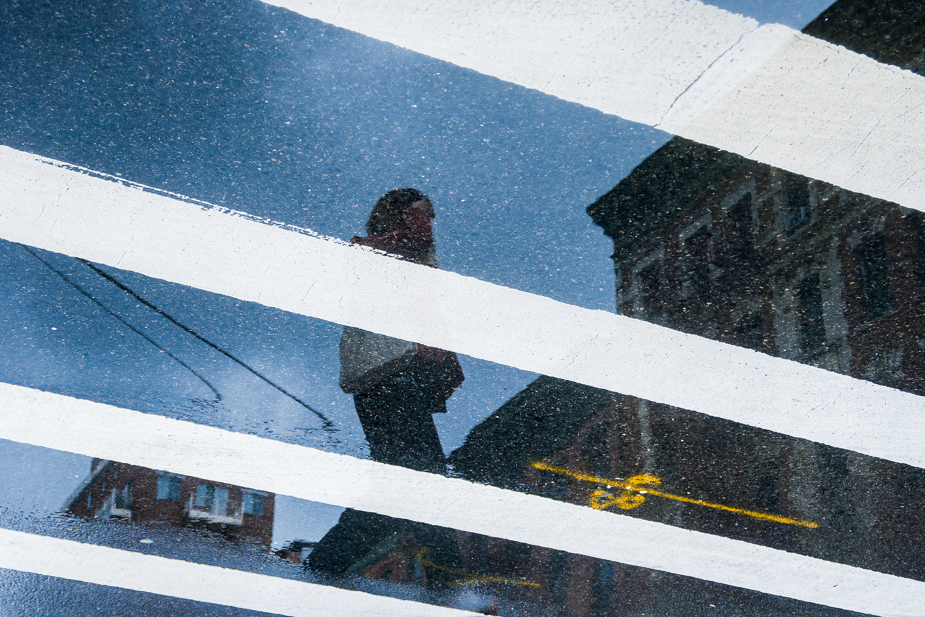 A street photograph of a pedestrian appearing in the reflection of wet pavement