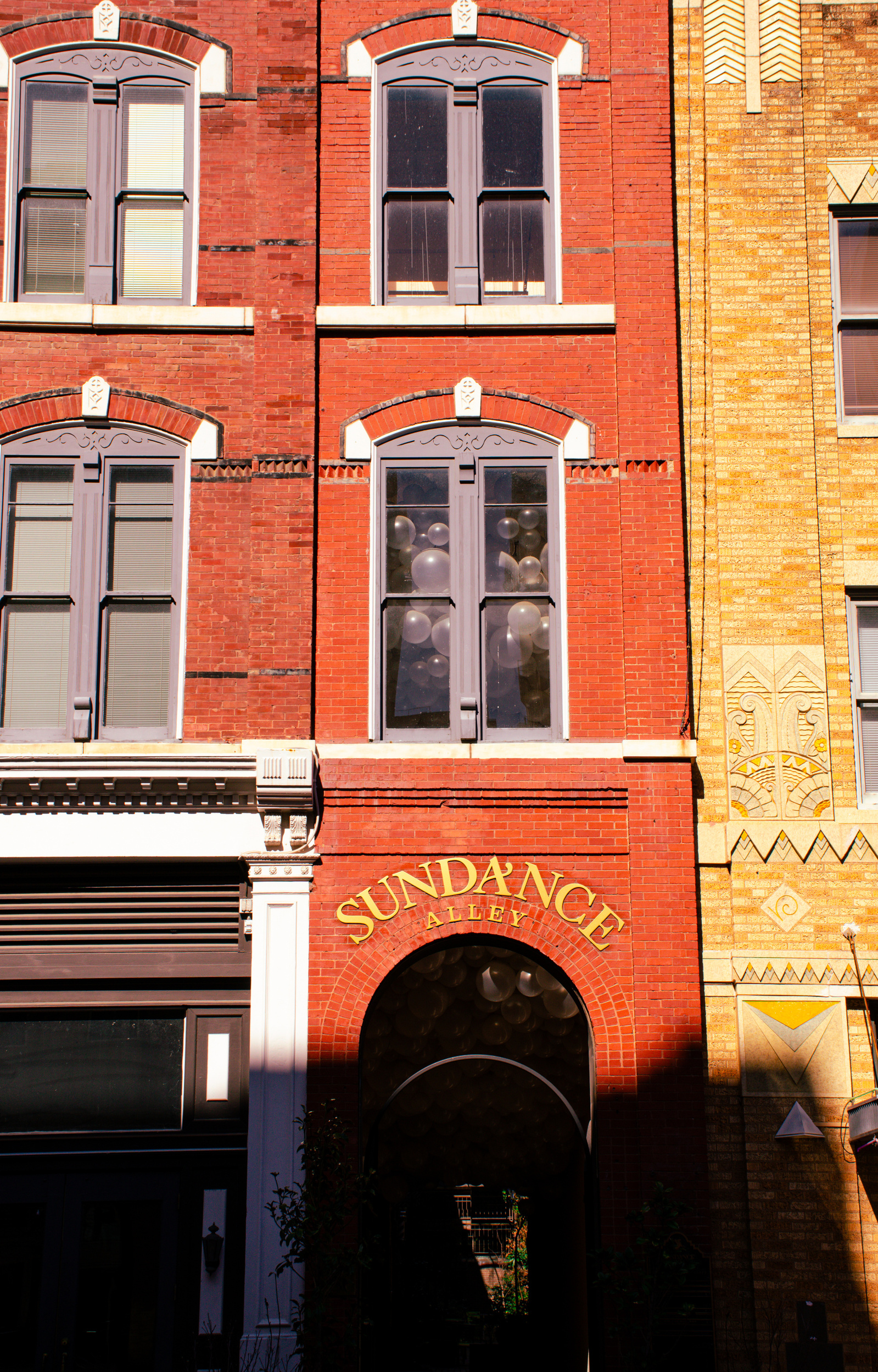 Balloons press against upstairs windows in this alleyway named Sundance. Above, red bricks and curved windows frame the surreal scene like a stage set.