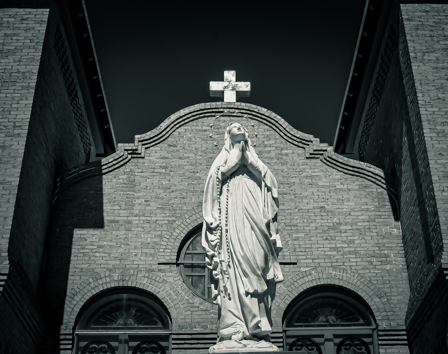 Close-up of a Virgin Mary statue, rosary in hand, cross above. Her gaze is cast upward, lips gently parted in silent pleading. Framed by the curve of old brick and the weight of shadow, she stands between earth and divine.