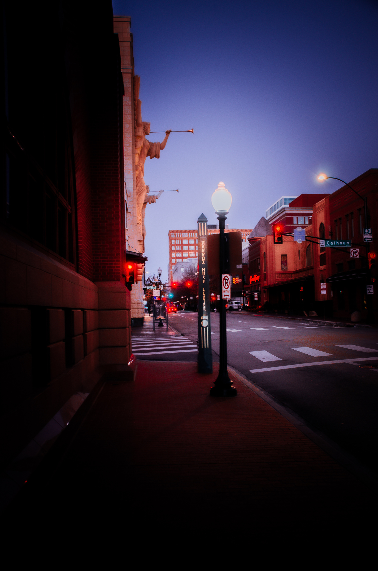 The intersection sits hushed under dawn or dusk. Streetlights glow against the sky while downtown waits to inhale.